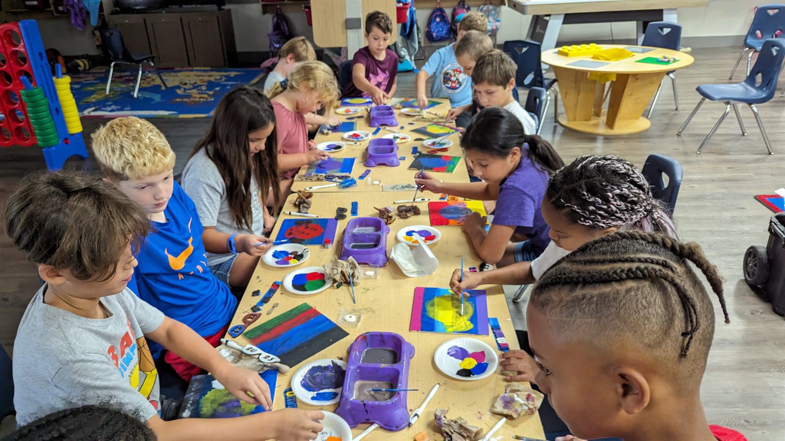 Kids seated around a long table painting colorful school group projects